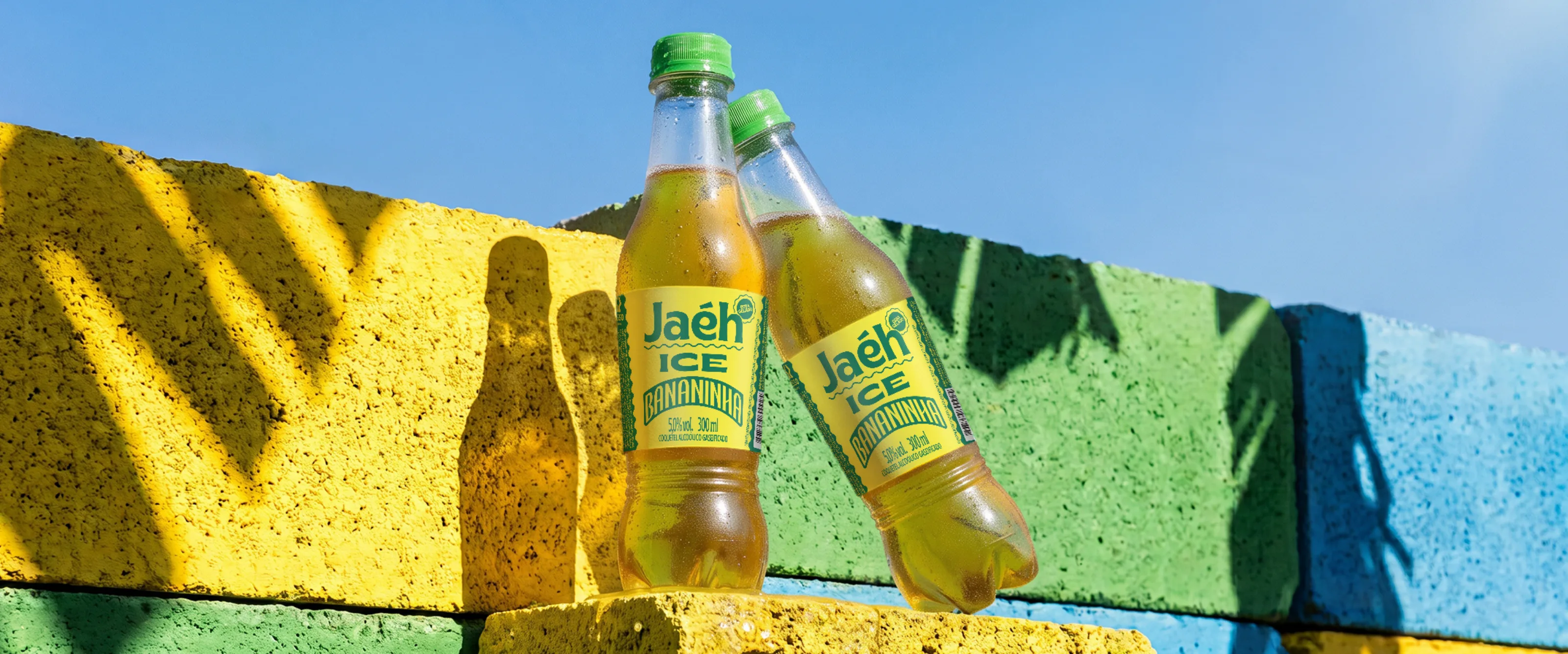 Garrafas geladas de Jaéh Ice Bananinha sobre blocos de concreto amarelos e verdes, ao ar livre.